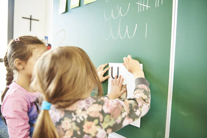 Zwei Schülerinnen hängen ein Blatt Papier an die Kreidetafel